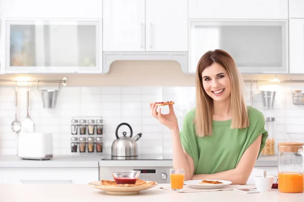 Beautiful woman eating tasty toasted bread with jam in kitchen