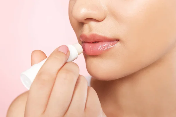 Young woman applying balm on her lips against color background, closeup