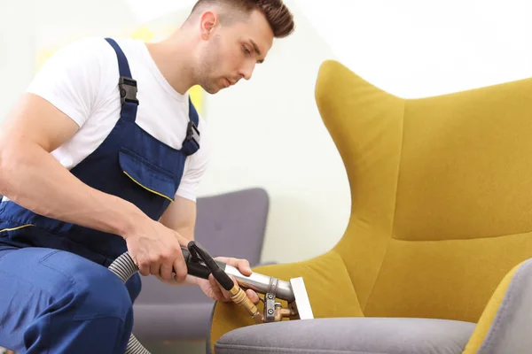 Male worker removing dirt from armchair with professional vacuum cleaner indoors