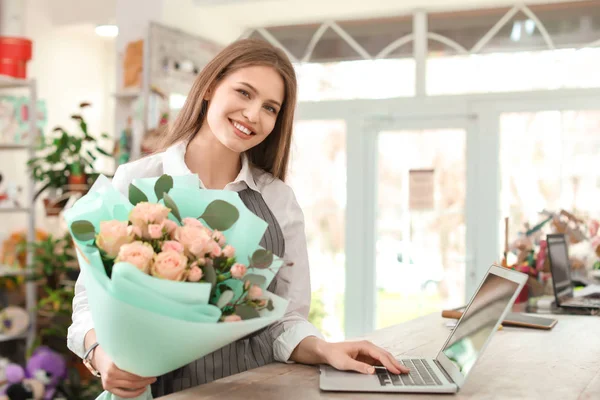 Female florist with bouquet and laptop working in flower shop
