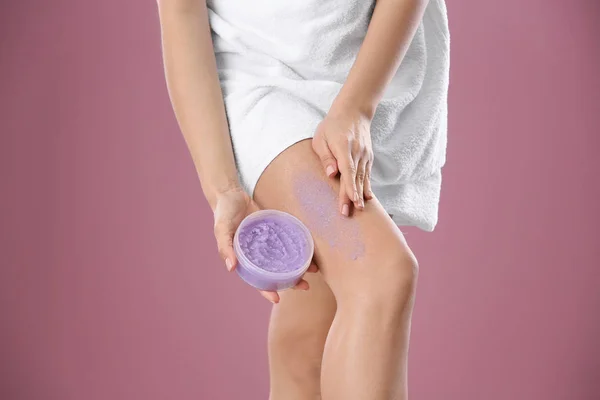 Young woman applying body scrub on leg against color background