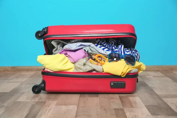 Open suitcase with different clothes and accessories packed for summer journey on floor near color wall