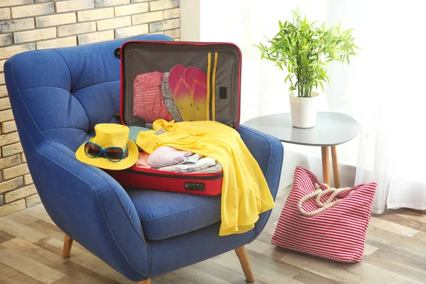 Open suitcase with different clothes and accessories for summer journey on armchair