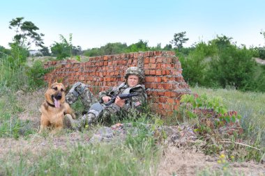 Askeri üniformalı bir adam ve Alman çoban köpeği kırık tuğla duvarın yanında.