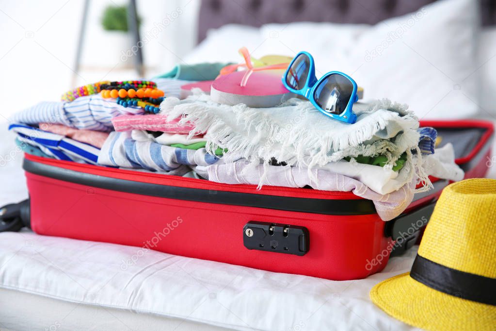 Open suitcase with different clothes and accessories for summer journey on bed