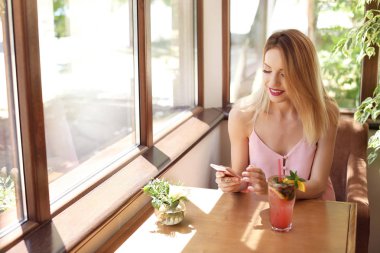 Kafede masada lezzetli doğal limonata yudumlarken cep telefonu kullanan genç kadın. Detoks içecek