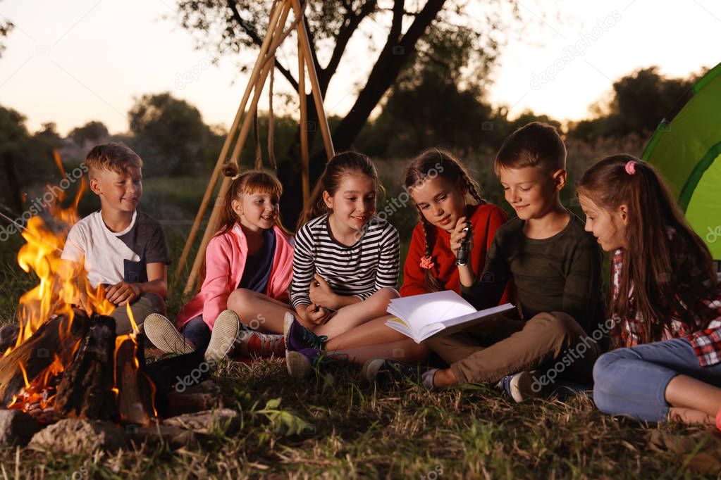 Little children reading book with flashlight outdoors. Summer camp