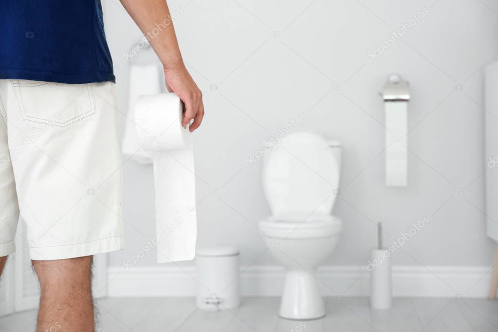 Young man with bath tissue standing near toilet bowl at home