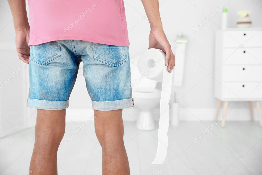Young man with bath tissue standing near toilet bowl at home