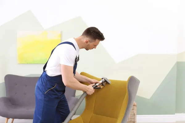 Male worker removing dirt from armchair with professional vacuum cleaner indoors