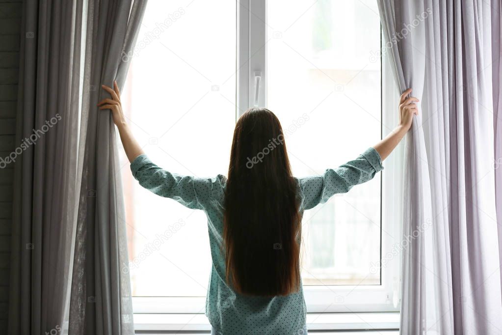 Young woman opening curtains in room near big beautiful window