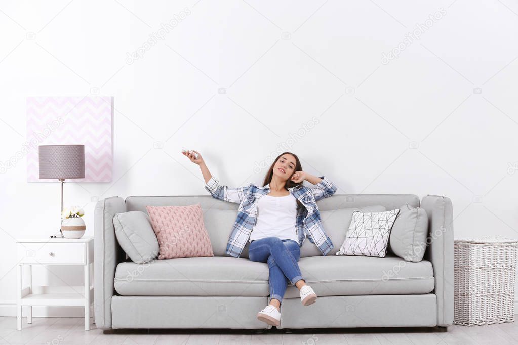 Young woman switching on air conditioner while sitting on sofa near white wall