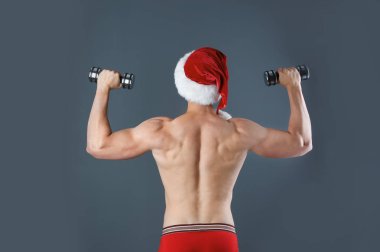 Noel Baba şapkası gri arka plan üzerinde dumbbells ile genç kaslı adam