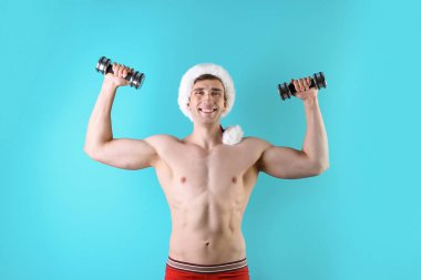 Noel Baba şapkası renk arka plan üzerinde dumbbells ile genç kaslı adam