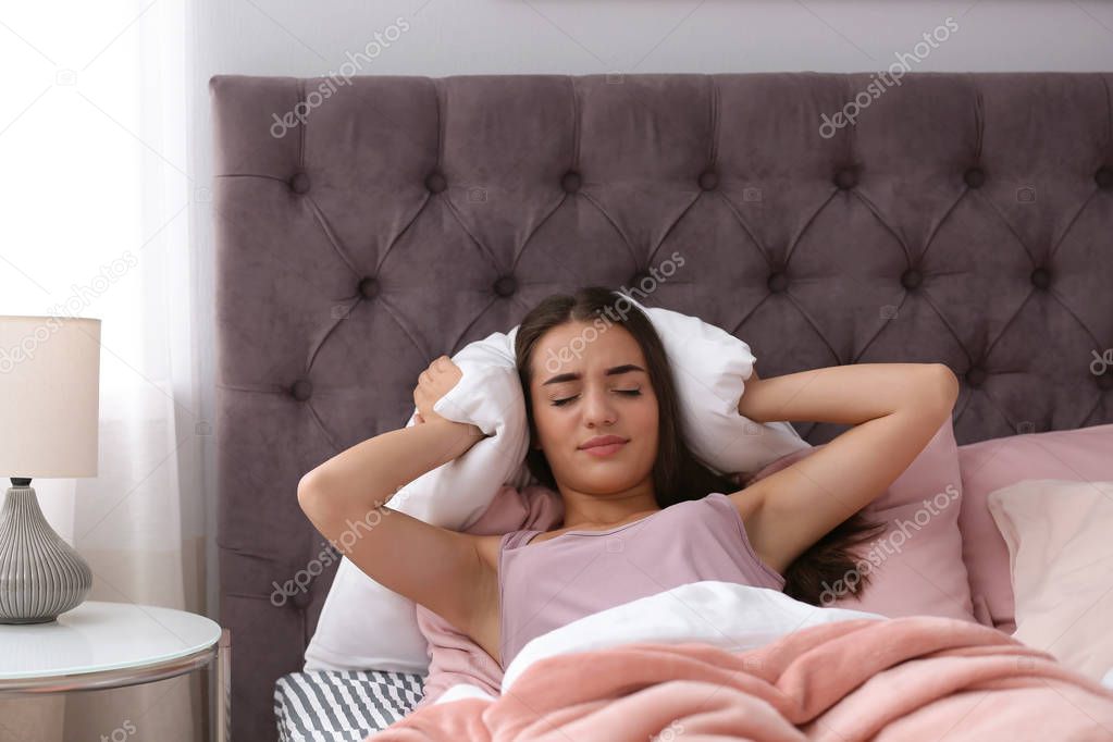 Young woman covering ears with pillow while trying to sleep in bed at home. Early morning