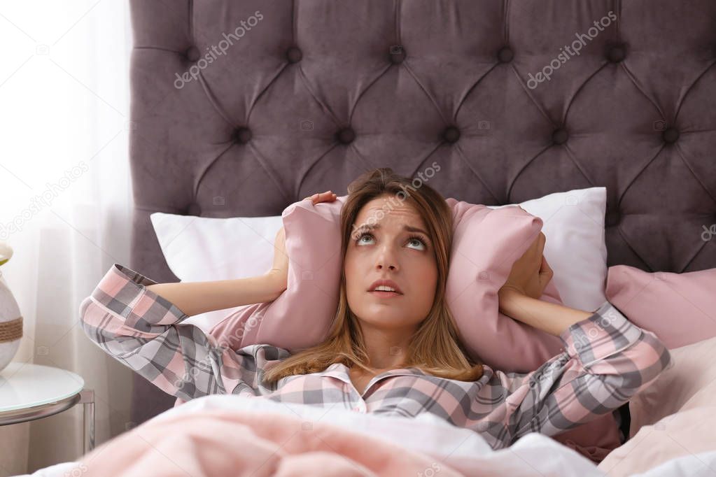 Young woman covering ears with pillow while trying to sleep in bed at home. Early morning