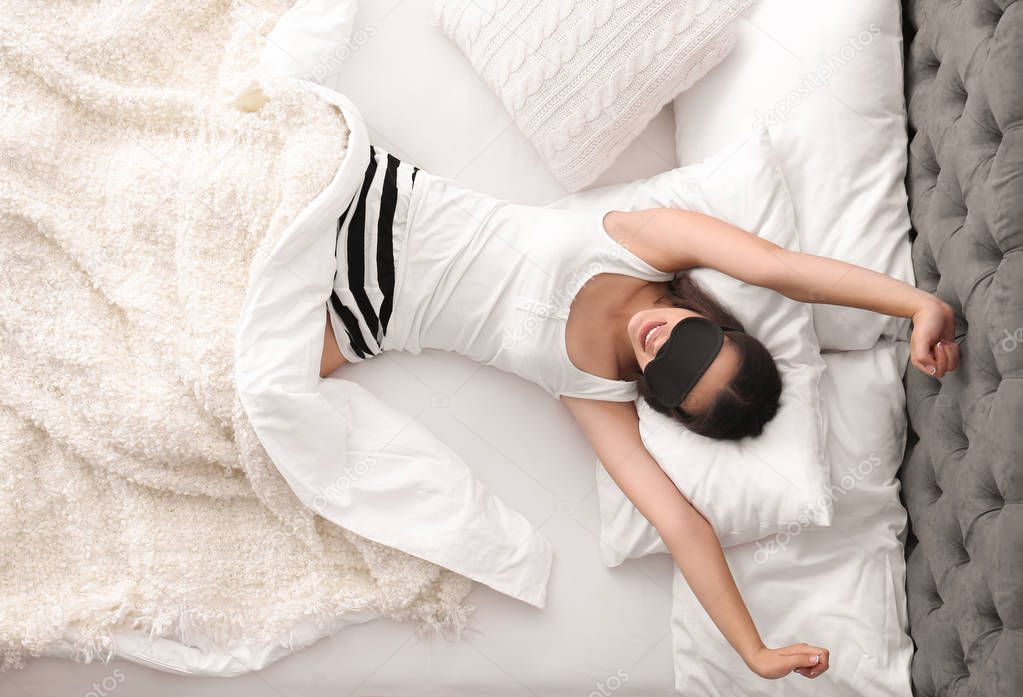 Young beautiful woman with sleeping mask stretching in bed at home, top view