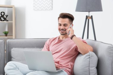 Evde kanepede dizüstü bilgisayar kullanırken cep telefonuyla konuşan adam