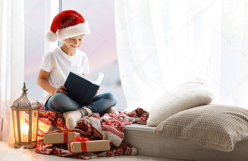 Cute little child in Santa hat reading Christmas story while sitting on windowsill at home