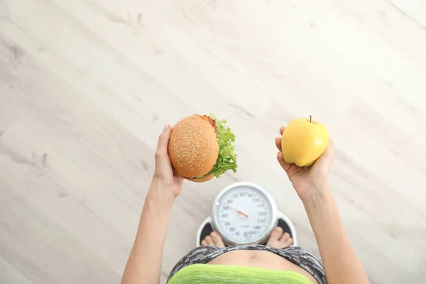 妇女拿着美味的三明治和新鲜的苹果 同时测量她的体重在地板上的鳞片 顶部的看法 饮食与不健康食品的选择 — 图库照片
