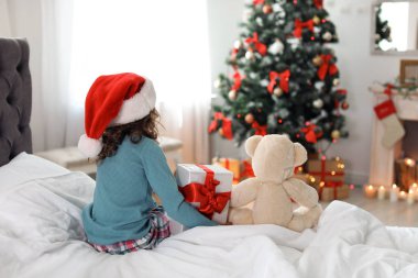 Sevimli küçük çocuk oyuncak ayı ve Noel hediye kutusu evde yatakta oturup Noel Baba şapkalı