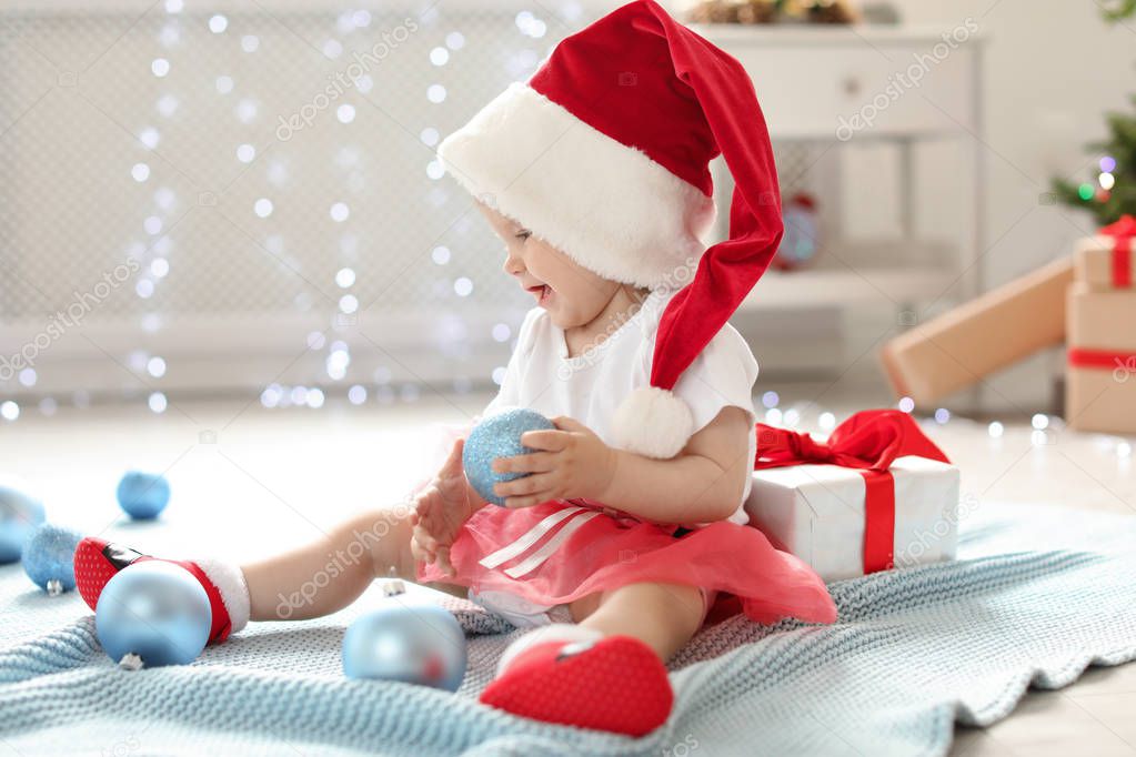 Cute baby in festive costume playing with Christmas decor on floor at home