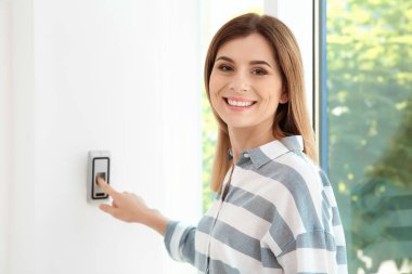 Young woman pressing fingerprint scanner on alarm system indoors clipart