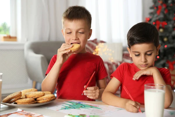 Little boy eating cookies while his friend drawing picture at home. Children celebrating Christmas
