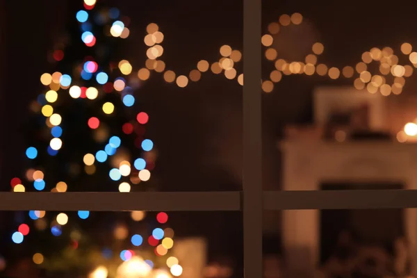 Blurred view of stylish living room interior with Christmas lights and fireplace at night through window
