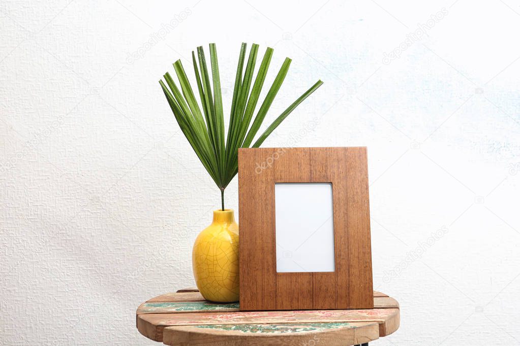 Blank frame and vase with tropical leaf on table near white wall. Mock up for design