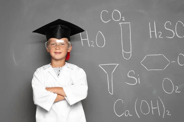 Little school child in laboratory uniform with graduate cap and chemical formulas on grey background