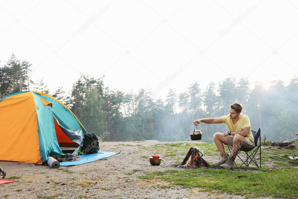 Young man holding kettle over bonfire near camping tent outdoors