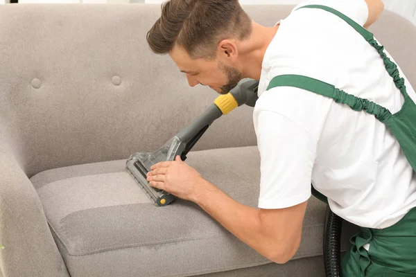 Male janitor removing dirt from sofa with upholstery cleaner indoors