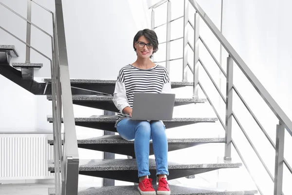Young woman with modern laptop sitting on stairs indoors
