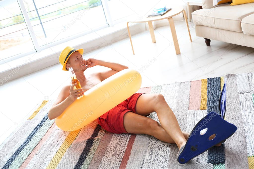 Shirtless man with inflatable ring and bottle of drink wearing flippers on floor at home