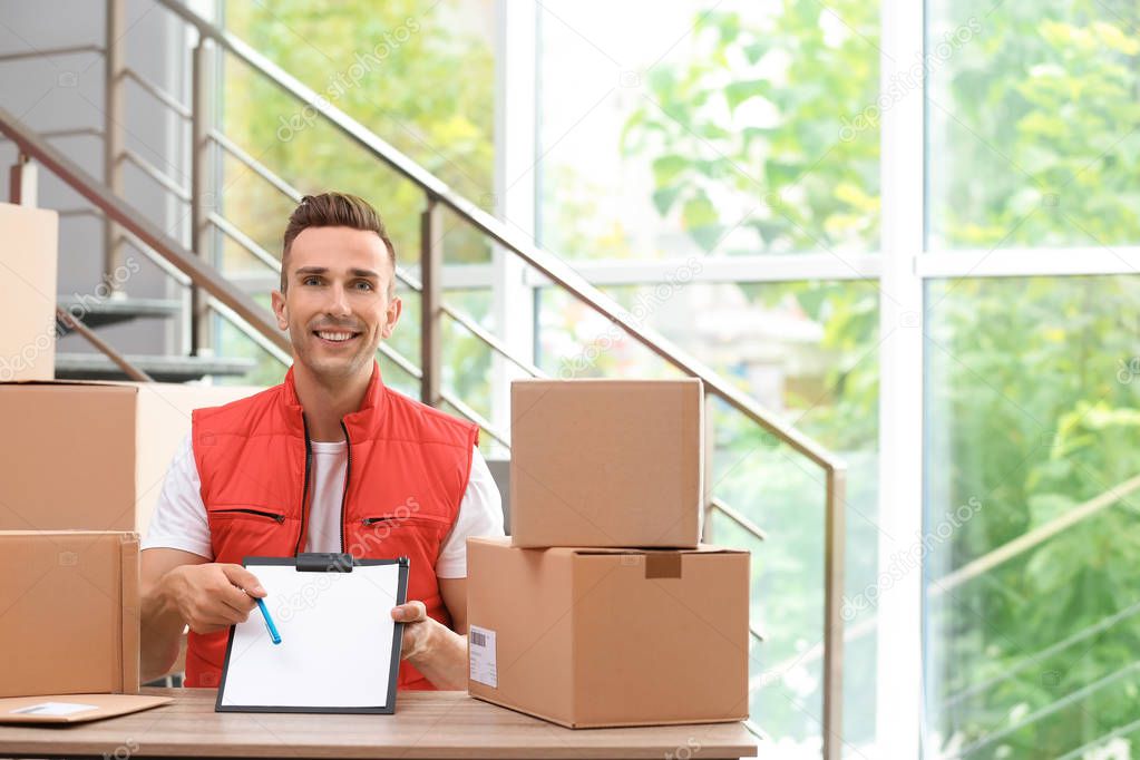 Young courier with clipboard among parcels at delivery department. Space for text