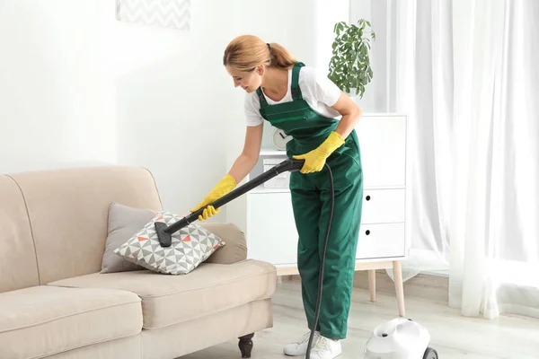 Female janitor removing dirt from sofa cushion with steam cleaner in room