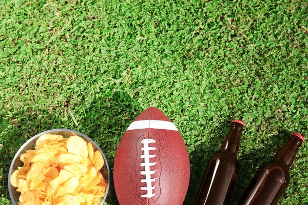 Ball for American football, beverage and chips on fresh green field grass, flat lay. Space for text