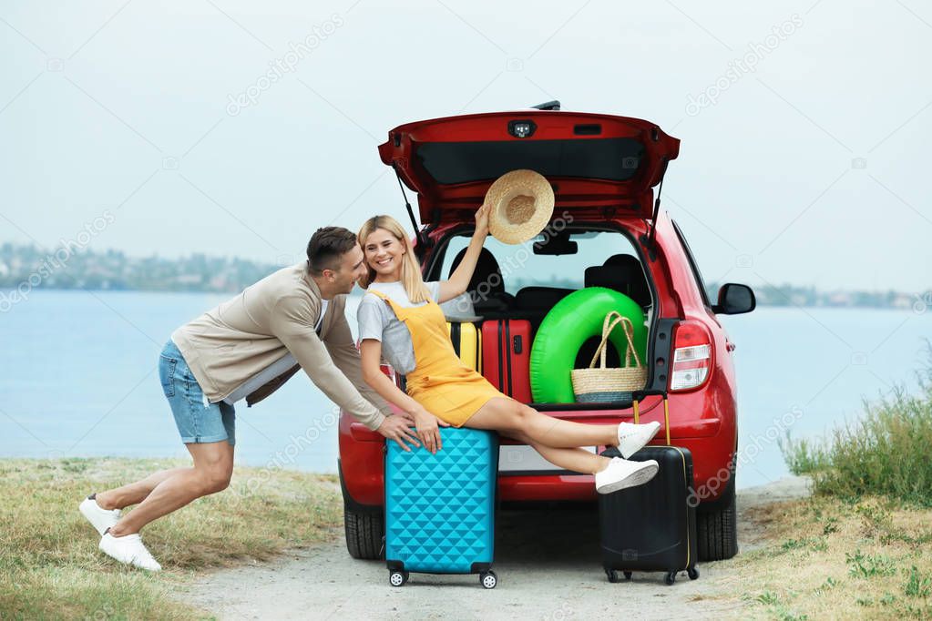 Couple having fun near car trunk with suitcases on riverside