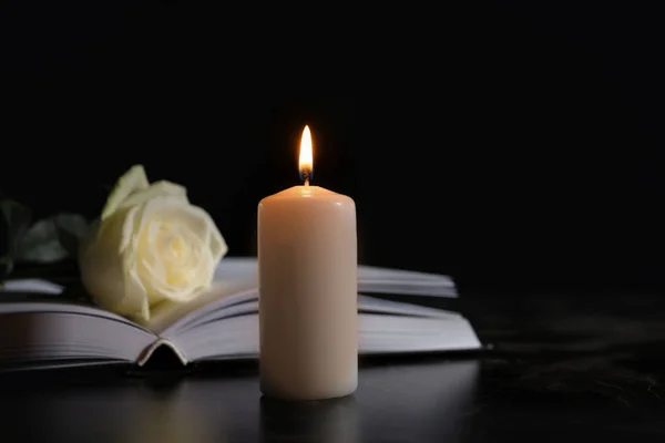 Burning candle, book and white rose on table in darkness, space for text. Funeral symbol