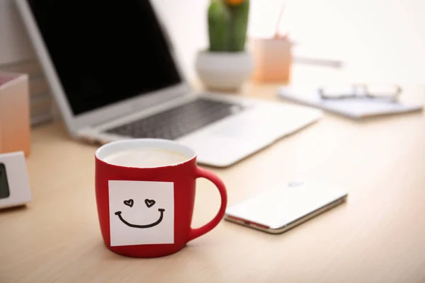 Sticky note with funny face attached to cup of coffee on office table. Space for text