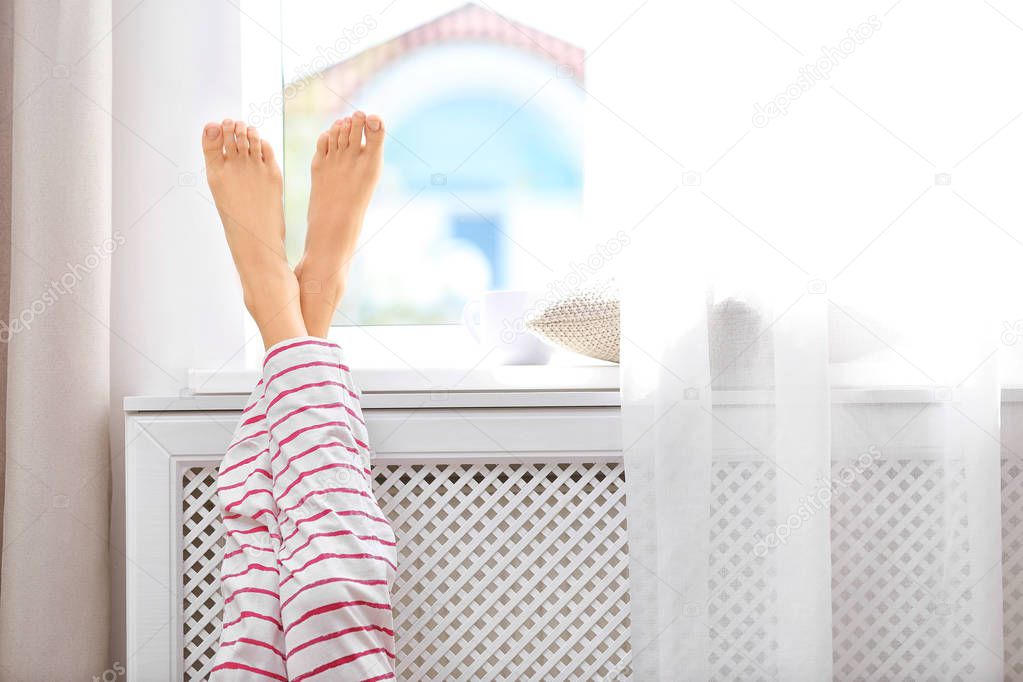 Woman warming up with feet on heater near window