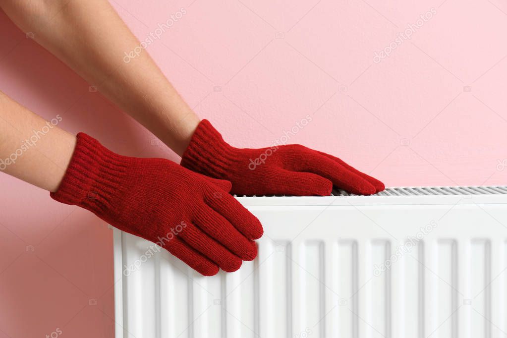 Woman in gloves warming hands on heating radiator near color wall
