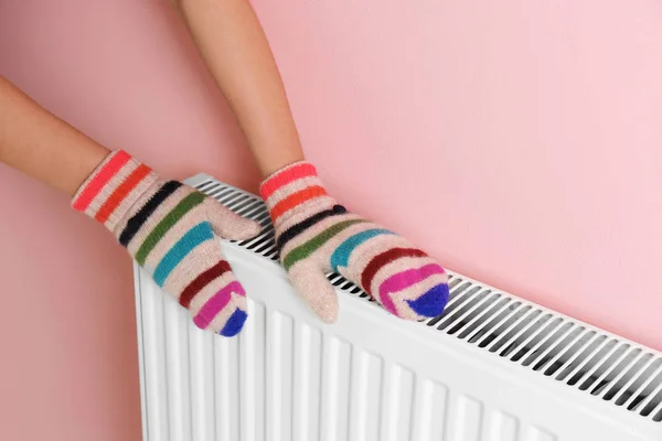 Woman in mittens warming hands on heating radiator near color wall