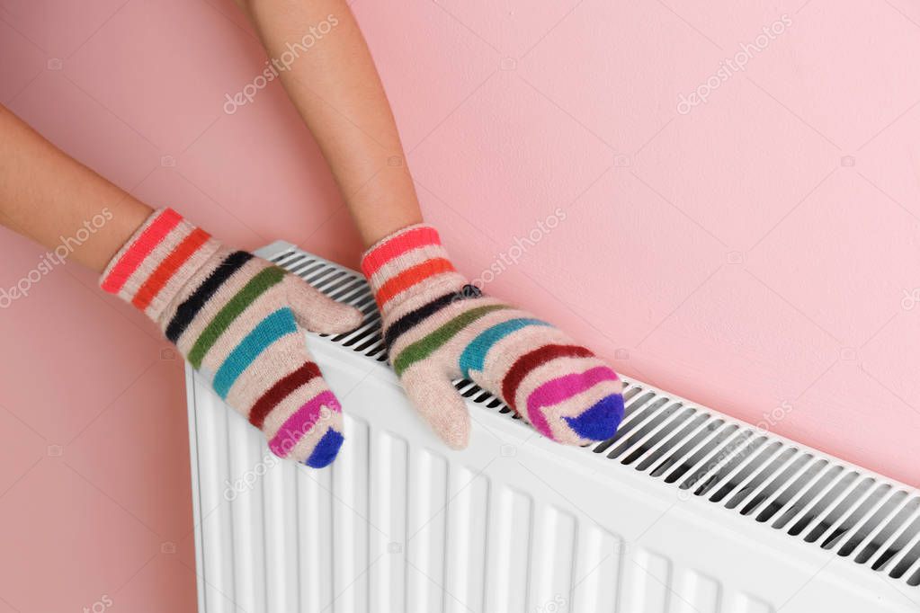 Woman in mittens warming hands on heating radiator near color wall