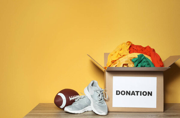 Donation box with clothes, shoes and rugby ball on table against color background. Space for text