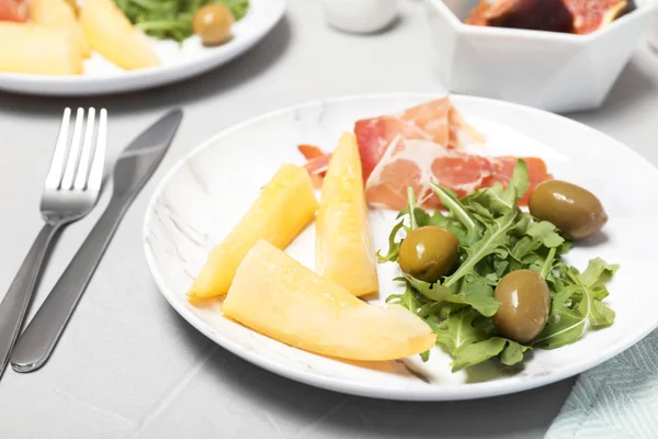 Plate with melon slices, prosciutto and olives on gray table