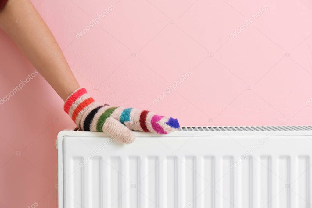 Woman in mitten warming hand on heating radiator near color wall. Space for text