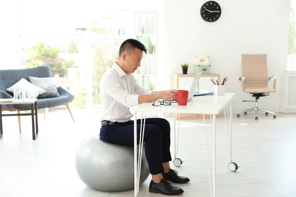 Young businessman sitting on fitness ball and using laptop in office. Workplace exercises
