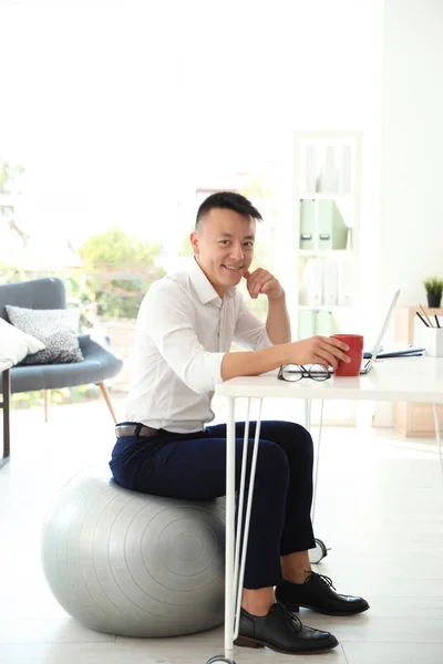 Young businessman sitting on fitness ball and using laptop in office. Workplace exercises